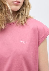 Közeli kép egy személyről, aki rózsaszín Pepe Jeans London pólót visel, szőke hullámos haja van, és az ajkak feletti arcvonások nem láthatók.