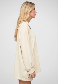 Beige oversized hettegenser med lange ermer, knappdetalj ved ermene og rundet nederkant, laget av myk, teksturert stoff.