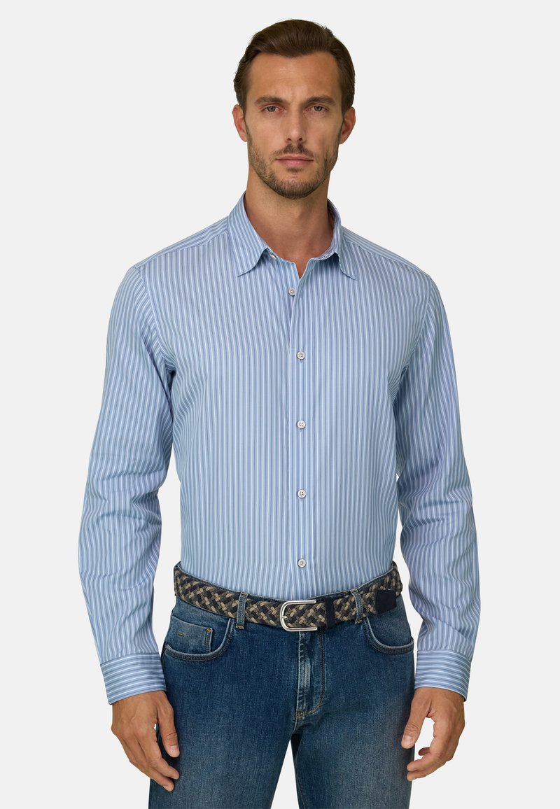 Homme portant une chemise boutonnée à rayures verticales bleu clair et blanc, rentrée dans un jean bleu avec une ceinture tissée, debout face à l'avant.