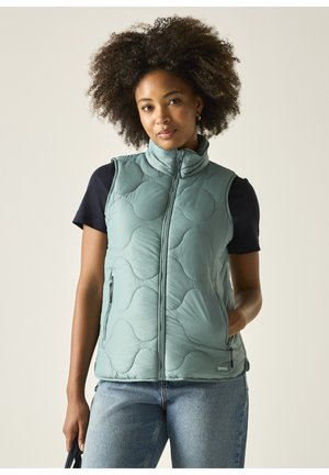 Femme aux cheveux bouclés portant une veste matelassée bleu clair sur un t-shirt noir à manches courtes et un jean bleu, debout avec une main dans la poche.