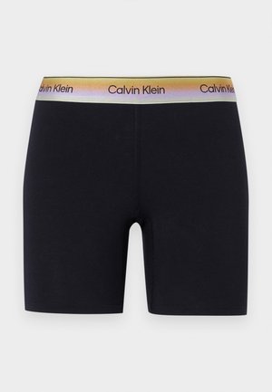 Mustad keha järgivad lühikesed püksid laia metallik-kuldses vöökohas, millel on korduvad "Calvin Klein" logod musta tekstiga.