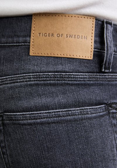 Jeans de mezclilla gris oscuro con una textura suave, que cuentan con una etiqueta de cuero marrón cosida en la parte trasera con "TIGER OF SWEDEN" grabado.
