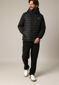 Homme barbu souriant, portant une veste matelassée noire avec capuche, un pantalon noir et des baskets blanches, marchant avec une main dans la poche.