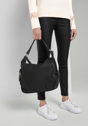 Sac à épaule synthétique noir avec deux poches zippées, texture lisse et une bandoulière amovible. Le modèle porte un pantalon skinny noir et des baskets blanches.