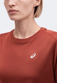 Gros plan d'une personne portant un maillot de sport couleur rouille avec un logo blanc ASICS sur la poitrine, montrant une partie du cou et du bas du visage.