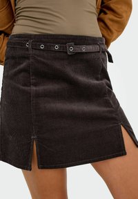 Gonna mini in corduroy marrone con passanti per cintura e una cintura abbinata, caratterizzata da fessure frontali su entrambi i lati, indossata con una maglietta e un giubbotto marrone chiaro.