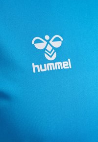 Blå idrottströja med en strukturerad tyg och en framträdande vit Hummel-logotyp, designad med en lekfull bi-motiv och geometriska detaljer.