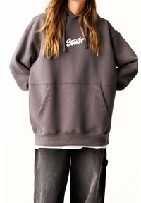 Personne portant un sweat à capuche gris surdimensionné avec poche avant et logo blanc, associé à un pantalon noir ample sur un fond uni.