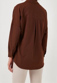 Donna che indossa una camicia lunga marrone a maniche lunghe, morbidamente aderente, con dettagli a pieghe sulla schiena, abbinata a pantaloni beige chiaro.