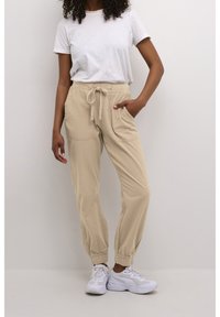 Beige Jogginghosen mit einem elastischen Bund, seitlichen Taschen und gerippten Bündchen, kombiniert mit einem schlichten weißen T-Shirt und weißen Sneakers.