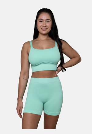 Junge Frau lächelt, trägt ein mintgrünes, geripptes Crop-Top und passende hochtaillierte Shorts, steht mit einer Hand in der Hüfte.