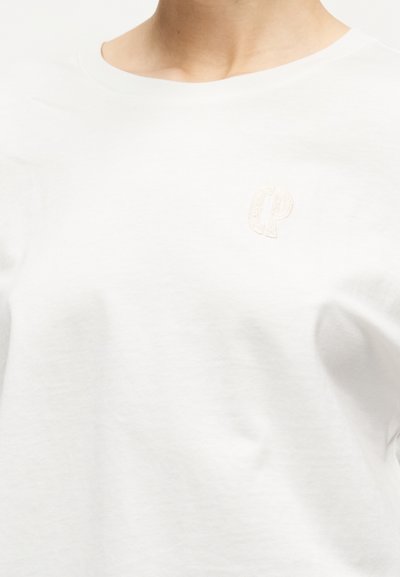 Άτομο που φοράει ένα απλό λευκό μπλουζάκι με στρογγυλή λαιμόκοψη και ένα διακριτικό κεντητό λογότυπο στο πάνω αριστερό μέρος του στήθους.