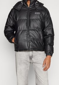 Czarna kurtka puchowa z błyszczącym wykończeniem, z zamkiem z przodu, regulowanym kapturem, elastycznymi mankietami i logo na piersi. Teksturowany, pikowany wzór.