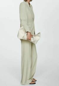 Persoon in een lichtbeige pak met wijde broek, die een grote bijpassende beige clutch vasthoudt, staande tegen een effen witte achtergrond.