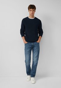 Dunkelblauer gerippter Pullover mit weißem Rundhalsausschnitt, kombiniert mit hellblauen Jeans und weißen Sneakern. Das Model steht aufrecht mit den Händen in den Taschen.