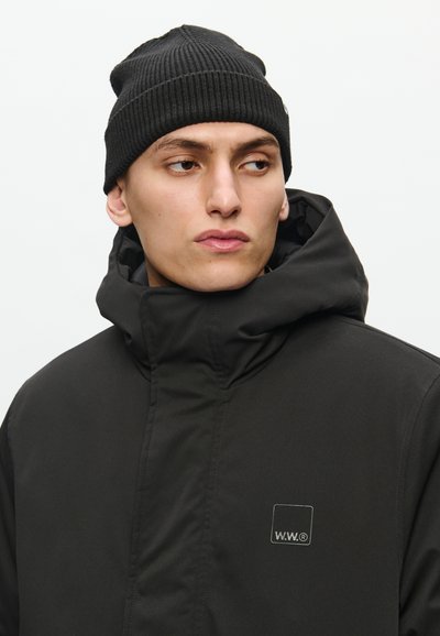 Black, padded jacket with high collar and logo patch, paired with a ribbed black beanie. Smooth fabric with lightweight texture.