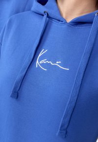 Modra bombažna kapuca z belim vezenim logotipom. Ima kapuco z vrvico in gladko teksturo.