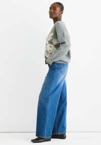 Gray knit sweater with floral embroidery in cream; paired with wide-leg blue denim jeans. Model wears black flats, stands in profile.