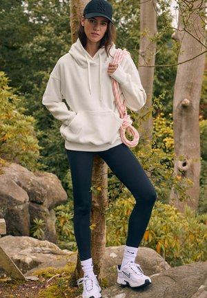 Sudadera blanca con cordones, leggings negros y zapatillas blancas. Lleva puesta una gorra negra y sostiene una cuerda rosa y blanca frente a un fondo natural.