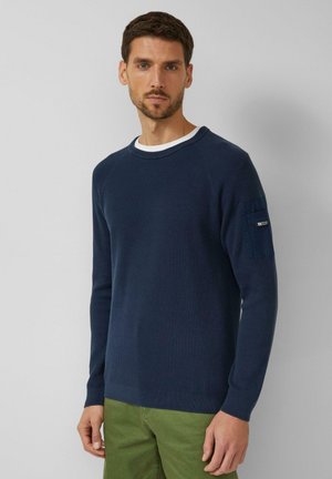 Marineblauer, gerippter Pullover mit Rundhalsausschnitt und langen Ärmeln; verfügt über eine aufgesetzte Tasche am linken Ärmel; kombiniert mit grünen Hosen.