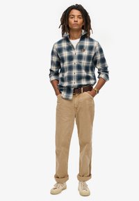 Camisa de franela a cuadros en azul y crema, abotonada y con las mangas arremangadas. Un pantalón de pana beige y zapatos beige completan el conjunto.