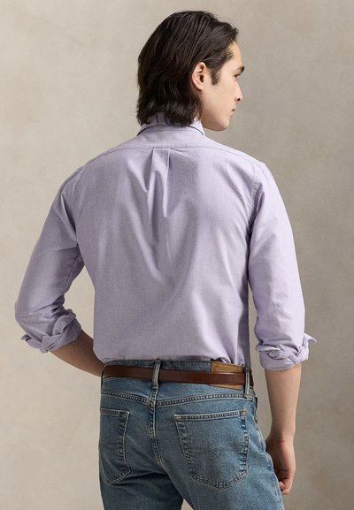 Ανοιχτό μωβ πουκάμισο με κουμπιά και γυρισμένα μανίκια, με σχεδιασμό yoke και πτυχή στην πλάτη, φορεμένο με μπλε τζιν και καφέ ζώνη.