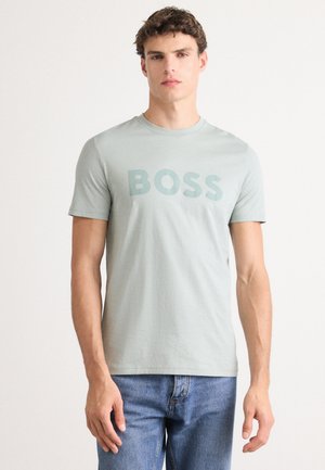 Hellgrünes Baumwoll-T-Shirt mit dem Wort "BOSS" in einer dunkleren Schattierung aufgedruckt, runder Ausschnitt und kurzen Ärmeln. Getragen mit blauen Denim-Jeans.