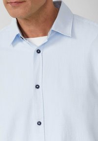 Camicia button-up di un blu chiaro, con texture, colletto rigido e bottoni scuri. Il colletto interno presenta un dettaglio in tessuto a contrasto.