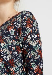 Sweter w kwiatowy wzór z granatową bazą, ozdobiony małymi czerwonymi, białymi i zielonymi liśćmi. Miękka tekstura i luźny krój z dekoltem w kształcie litery V.