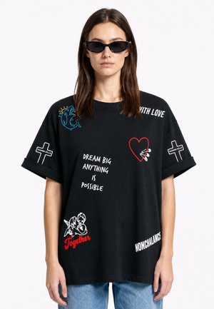 Vrouw met lang kastanjebruin haar die zwarte cat-eye zonnebrillen draagt en een zwart oversized T-shirt met diverse witte en rode grafische teksten en afbeeldingen.
