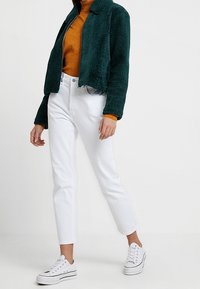 Grøn textured cropped jakke over en orange sweater, parret med hvide straight-leg jeans og hvide sneakers. Bløde materialer, afslappet design.