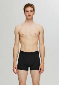 Νεαρός άνδρας στέκεται, κοιτάζοντας μπροστά, φορώντας μαύρα μποξεράκια μπροστά σε απλή ανοιχτή γκρι φόντο.