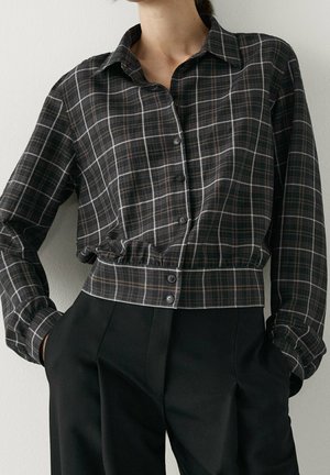 Femme portant une chemise à carreaux foncés boutonnée, rentrée dans un pantalon noir taille haute, les mains dans les poches, debout contre un mur blanc.