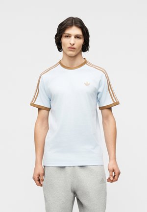 Fiatal férfi középhosszú sötét hajjal, világoskék Adidas pólót visel barna csíkokkal és világosszürke Adidas melegítőnadrágot, állva.