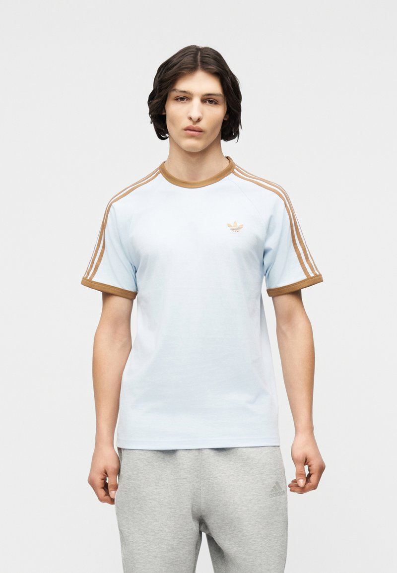 Νέος άνδρας με μεσαίου μήκους σκούρα μαλλιά, φορώντας ανοιχτό μπλε μπλουζάκι Adidas με καφέ ρίγες και ανοιχτό γκρι παντελόνι φόρμας Adidas, όρθιος.