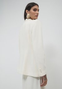 Donna con capelli scuri che indossa un blazer bianco, grandi orecchini rotondi dorati e pantaloni bianchi, girata leggermente di spalle rispetto alla fotocamera su uno sfondo semplice.