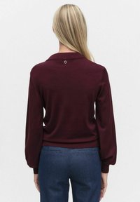 Burgundzkie sweter z kołnierzykiem polo, luźnymi rękawami i okrągłym detalem z tyłu. W połączeniu z dopasowanymi niebieskimi spodniami.