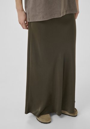 Persona che indossa una lunga gonna verde oliva, scarpe beige in stile mocassino e una t-shirt oversize color taupe, in piedi contro uno sfondo neutro.