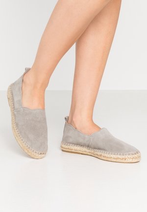 Jalat yang mengenakan sepatu espadrille suede abu-abu muda dengan sol yang dibalut jute, satu kaki sedikit diangkat di atas latar belakang lantai putih.