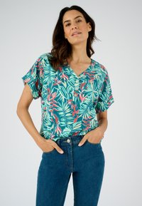 Blusa de manga corta con un patrón de hojas tropicales verdes; presenta un escote en V y cierre de botones. Combinada con jeans azules.