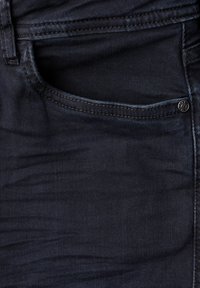 Tela de mezclilla azul oscuro con una textura suave, que presenta un bolsillo delantero, costuras visibles y un pequeño detalle de botón metálico.