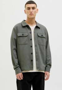 Gray cotton shirt jacket with a collar, two chest pockets, and black buttons, worn over a cream shirt with dark jeans. Textured fabric finish.