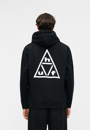 Osoba odjevena u crnu majicu s kapuljačom s velikim bijelim geometrijskim trokutastim uzorkom i slovima "h", "u" i "f" na leđima, okrenuta leđima.