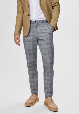 Homme portant un blazer marron clair, une chemise blanche, un pantalon gris à carreaux et des chaussures en daim beige, debout devant un fond clair.