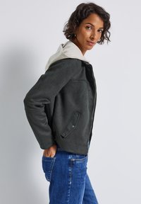Vrouw met kort krullend haar, gekleed in een donkergrijze zip-up jas over een beige hoodie en blauwe jeans, poseert met één hand in haar zak.