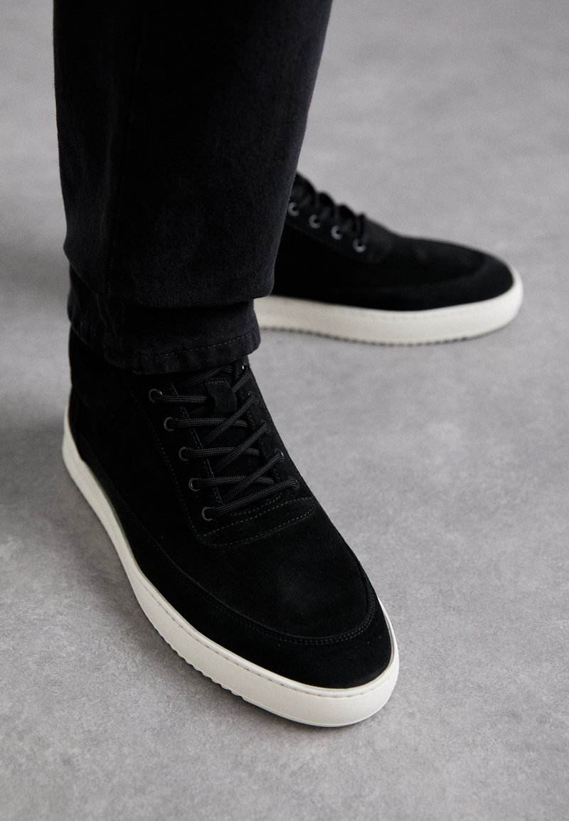 Sorte semskede høyhælte sneakers med hvit gummisåle, med snøring og sømdetaljer, plassert mot en nøytral bakgrunn.