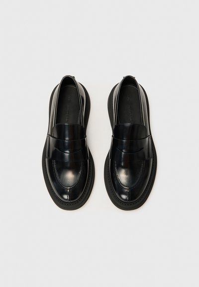 Mocassins en cuir noir brillant avec un bout arrondi, une large bande sur le devant, des coutures blanches et une semelle en caoutchouc noire épaisse.