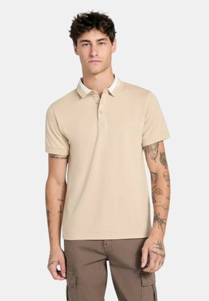 Giovane uomo con tatuaggi indossa una polo beige a maniche corte e pantaloni cargo marroni, in piedi davanti a uno sfondo chiaro e uniforme.