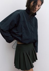 Kvinde med mørkt bølget hår iført en løs sort jakke og en kort mørkegrøn plisseret nederdel mod en ensfarvet lys baggrund.