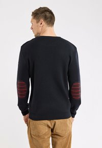 Mann trägt einen schwarzen Pullover mit roten Streifen an den Ellenbogen und braunen Cordhosen, mit dem Rücken zur Kamera vor einem schlichten weißen Hintergrund.